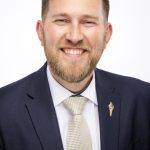Headshot of a smiling man with short hair and a beard wearing a navy suit, white shirt, and yellow tie, against a light background