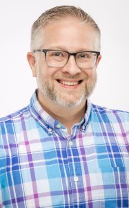 Headshot of a man with short gray hair, glasses, and a trimmed beard, wearing a blue and purple plaid button-up shirt, smiling against a white background