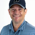 Portrait of a smiling Bushnell University staff member wearing a Bushnell baseball cap and blue striped polo shirt against a light background.