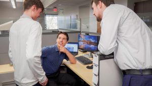 Students engage in professional workplace discussions while working in an office environment equipped with computers and collaborative workspaces.