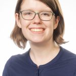 Headshot of a woman with short light brown hair and green-framed glasses, wearing a navy Bushnell University shirt, smiling against a white background