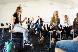 A group of students, some sitting on classroom desks and others standing nearby, casually talking.