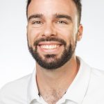 Headshot of a smiling man with short dark curly hair and a beard wearing a white collared shirt against a white background