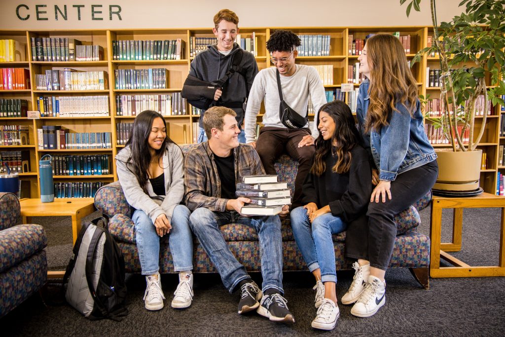 male and female students sitting on and around a couch in the library, stack of books on the lap of the middle student