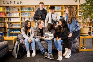 male and female students sitting on and around a couch in the library, stack of books on the lap of the middle student