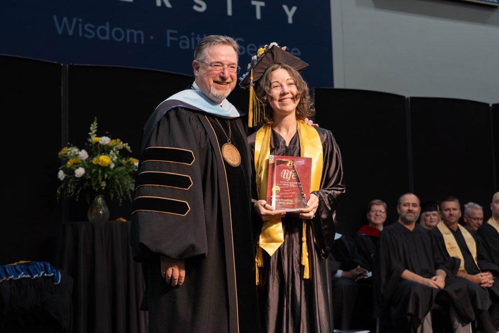 University president standing with a graduate in cap and gown holding an award plaque during a graduation ceremony