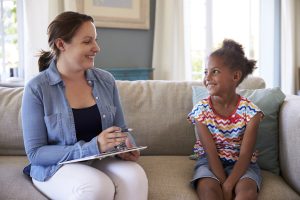 Child counselor sits with child client on a couch with clipboard in hand