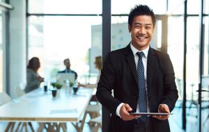 A young, confident businessman in professional clothing smiles while holding a tablet