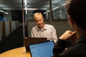 A student intern in a business meeting meets with another business intern while using their laptops