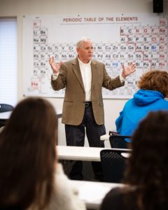 Male professor lectures at the front of class in brown jacket