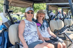 Two women smile while sitting in a golf cart during a sunny day at Pine Ridge Golf Club.