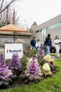 Prospective students walk past a Bushnell University campus sign and landscaped garden during a campus visit.